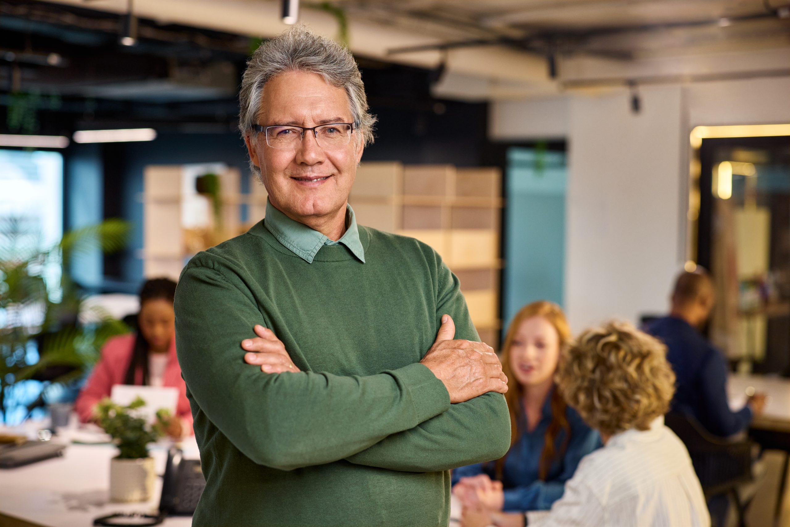 Portrait of reliable mature business man standing in modern office while his teamwork working in background. Successful mature entrepreneur in casual outfit looking at camera with satisfaction. Confident old man with eyeglasses standing with crossed arms with copy space.