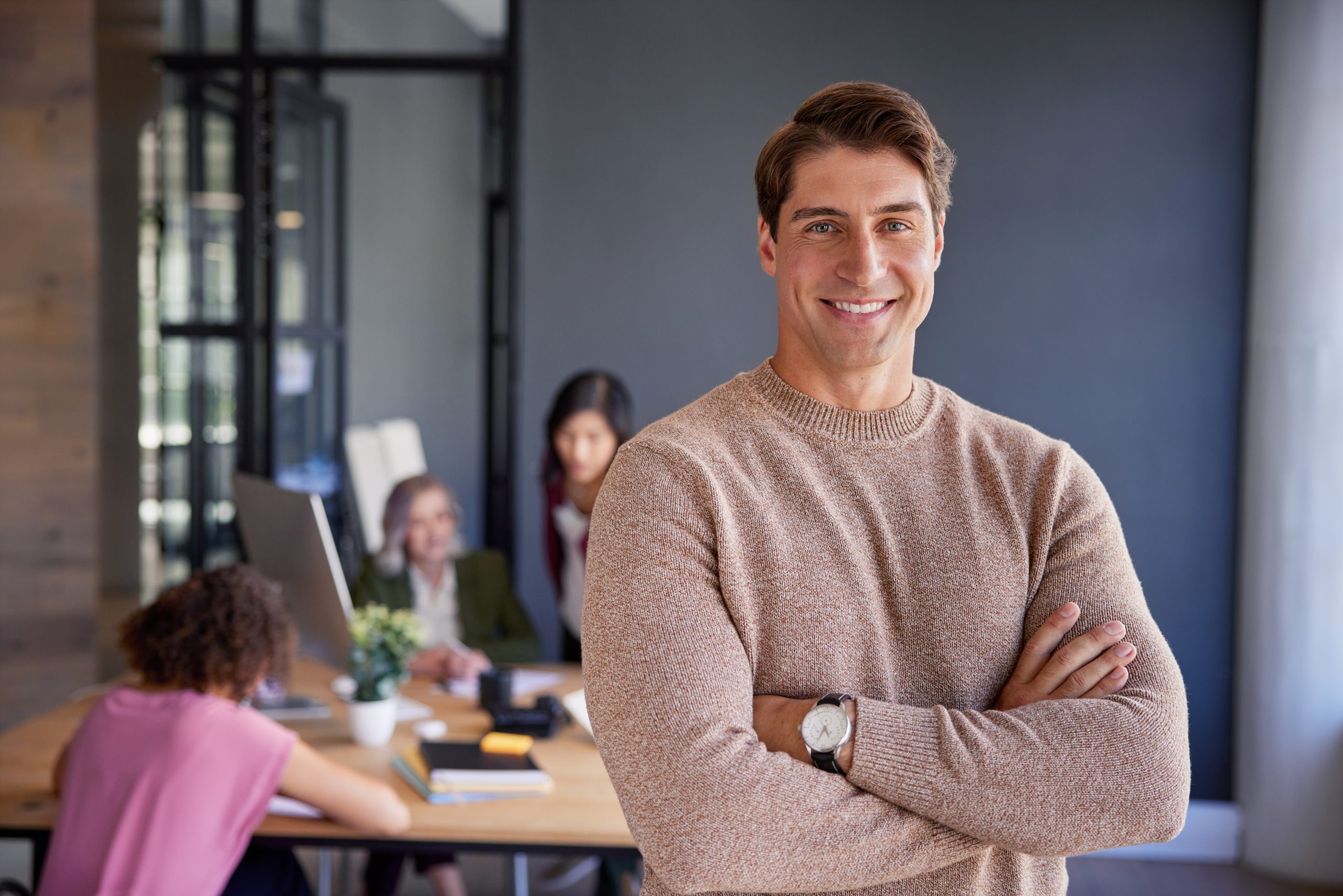 Successful mid adult businessman standing in front of a team with crossed arms. Smiling casual man looking at camera with a big smile while colleagues collaborating in the background. Portrait of reliable and confident business man in a creative office.