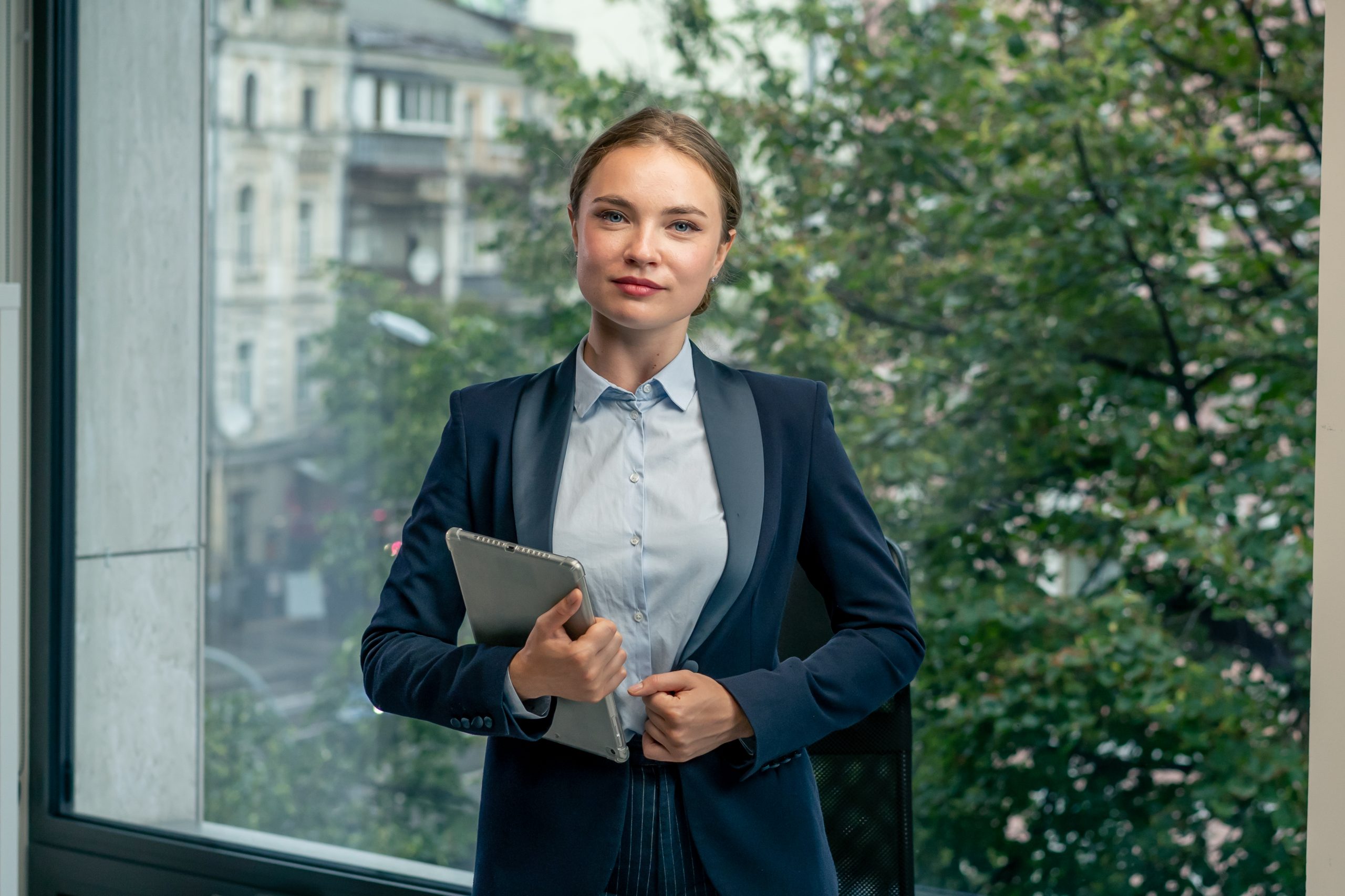 in a modern office with a large window a beautiful female worker stands in a blue shirt and a blue suit with a laptop and a tablet in her hand looking at the camera young boss